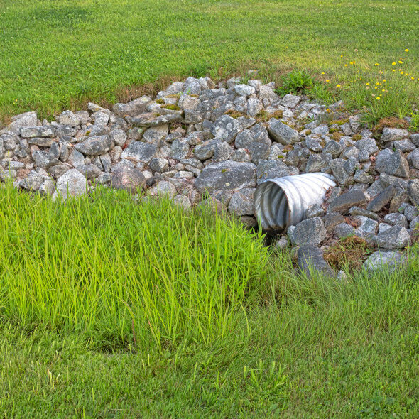 A stainless steel drainage culvert surrounded by rocks and green grass.  Effective drainage will have huge impacts on the health of your lawn and landscape.